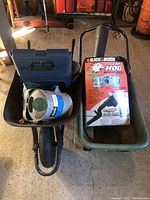 Two wheelbarrows side by side, one black plastic single-wheel and one green CSI plastic dual-wheel, containing assorted tools.