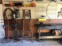 Photo showing entire setup with ladder, drill press, work bench, vice and wrenches, with electrical cords on pegboard