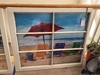 Large vintage wood window frame with 6 panes holding a beach scene poster of umbrella and three colorful beach chairs on sand and ocean background.