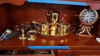 Photo of brass-colored tea serving set with pitcher, sugar bowl, creamer, and tray, two brass candle holders, and nautical clock on wooden stand