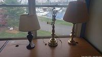 Three desk lamps are placed on a wooden desk by a window. Two lamps have pleated cream shades; one is black ceramic, the other brass. The third is a brass lamp without shade with visible wiring and switch.