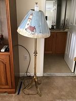 Floor lamp with brass tripod base standing upright with plastic-wrapped lighthouse-themed lampshade