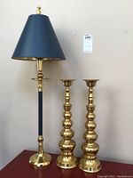 Table lamp with navy blue shade and gold metal base, next to two tall brass candle holders with bulbous design