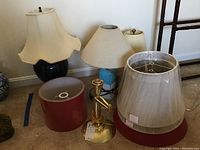 Four lamps with bases of various materials and designs, four assorted lampshades, and a small electric burner grouped on floor against wall.