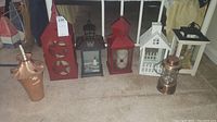 Photo showing seven lanterns and one copper oil lamp on the floor, varying colors and sizes, some designed for LED or candle use.