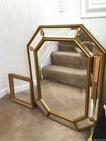 Two framed mirrors leaning against a wall by the stairs, showing the gold octagonal framed mirror and smaller rectangular mirror on carpeted floor.