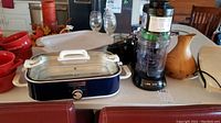 Countertop with slow cooker (blue base, glass lid), Ninja Spiralizer with attachments, red bowls, and other kitchenware.