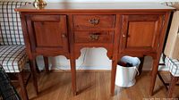 Front view of Pennsylvania House wooden buffet server with two side cabinet doors, two center drawers with brass handles, and scalloped apron detail, placed on hardwood floor between two patterned chairs.