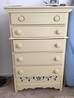 Front view of cream colored five drawer dresser with round knobs and blue floral detail on bottom drawer.