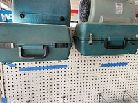 Two green hard plastic power tool cases stacked on pegboard shelf