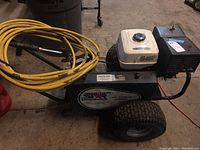 Side view of blue steel frame pressure washer with white Honda engine cover, black pump housing, pneumatic wheels and coiled yellow hose