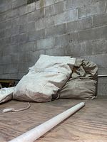 Two folded canvas tents in off-white and beige color resting on a wooden surface against a cinderblock wall. A white pole lies in front.