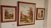 Three framed original landscape paintings depicting cottages and autumn trees on a wall.