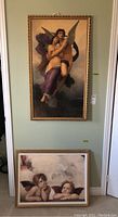 Photo showing two framed prints on board, one large vertical image of winged figures embracing and one smaller horizontal print of two cherubic angels resting on their arms.