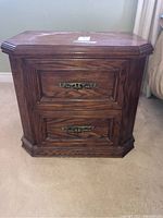 Front view of a carved wood bedside table with two drawers and ornate brass handles.