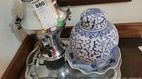 Photo showing silver-tone coffee carafe with handles, blue and white ceramic ginger jar on scalloped plate, and silver tray
