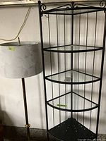 Black metal corner shelving next to brass floor lamp with fabric lampshade in a room with concrete floor and white wall.