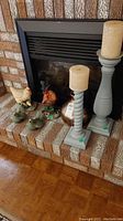 Overall view of two rooster figurines, two green bird figurines, the ceramic vase, and two gray candle holders with pillar candles on brick hearth