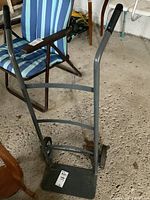 Front and side view of small metal dolly hand truck showing base plate, frame, wheels, and handle grips in a storage room.