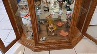 Glass display cabinet with wooden frame showing two shelves holding figurines, Royal Doulton clock, miniature tea cups, glass vases and goblets.