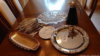 Full lot on wooden dining table showing flatware pieces, divided glass winter theme platter, marble cheese board, silverplate tray, tall black water pitcher with silver spout, and rectangular metal serving tray.
