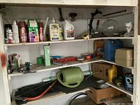 Photo of garage shelves showing various household products like crack filler, cleaners, sprays, and tools including a sprinkler, staplers, and a hacksaw