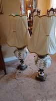 View of two tall table lamps with beige pleated fabric shades and decorative metal and glass bases.