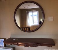 Wall-mounted painted wood hall table shelf with carved detailing below it and round matching mirror above, reflected window and light switch visible.
