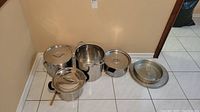 Four stainless steel pots with lids arranged on floor, along with stack of three metal baking pans/trays to right