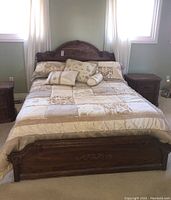 Front view of queen size bed frame with beige and cream patchwork comforter and multiple pillows, showing carved footboard.