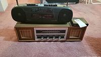 Black portable radio with double cassette player placed on top of a classic wood panel radio with analog dials and speaker grilles.
