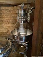 Silver plated coffee server with glass upper part, silver plated base and detailed silver handle with spout, resting on decorative holder