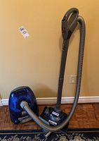 Blue Eureka vacuum cleaner with grey hose and floor head on wood floor against wall.