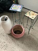 Photo showing two metal framed plant stands with decorative ceramic tile tops, two ceramic vases including one tall off-white vase containing an umbrella and shoe horn, and one pink round plant pot.