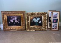 Four framed floral artworks arranged on beige carpet against wall. Two larger gold ornate framed oil paintings on the left, two smaller framed floral prints on the right grouped together.
