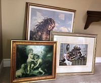 Three framed prints grouped on floor including a print of a woman with flowing hair, rustic family scene, and cherubic children print