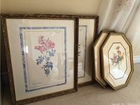 Four framed floral prints shown leaning against wall, two in ornate rectangular frames with floral pink flowers and decorative mats, one octagonal gold frame with bouquet print, another rectangular print partially visible