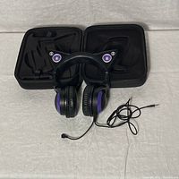 Wired purple cat ear headphones with microphone, attached cable with 3.5mm jack, and open hard carrying case behind them.