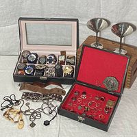 Black faux-leather watch box holding 11 wristwatches, adjacent red jewelry box, leather cuffs, necklaces, two metal goblets