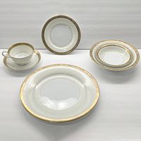 Group photo showing tea cup on saucer, bread plate, dessert bowl, and dinner plate all with gold rim detailing on white bone china.