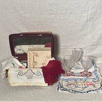 Vintage maroon Samsonite make-up case open showing interior mirror, hand embroidered linens with floral motifs, an unused pressed flower notebook with butterfly design, red and pink crocheted linens, and three clear etched glass goblets on embroidered linens.