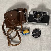 Yashica Lynx-1000 camera with lens cap, brown leather case, and roll of Ektachrome 100 film on cloth background.