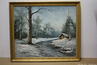 Front view of the framed oil painting depicting a winter scene with snow, trees, a river, and a cabin, signed by Chris Wells.