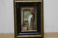 Framed print showing a priest wearing white robes standing at an open window, holding a book and rosary, looking out to a landscape of trees and hills.
