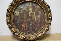 Front side of the round wall art showing a detailed historical scene with castle and figures, ornate gold frame with leaf and berry decorations