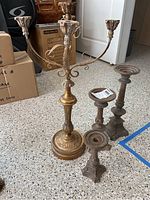 Full view of three wrought iron candleholders and one three-armed candelabra with antique hand finish, placed on a terrazzo floor next to cardboard boxes.