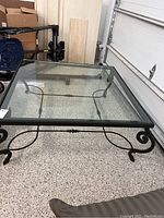 Full view of the square coffee table with clear glass top and decorative wrought iron base on a speckled floor.