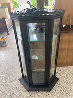 Full view of the black metal hexagonal curio cabinet with glass panels all around and ornate top edges.