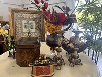 Overview of floral arrangement in glass vase, wooden decorative box, hand painted ceramic box and candle holders on table