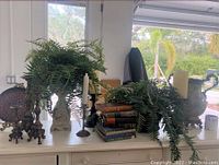 Wide view of white cabinet top showing three faux fern plants in vases, stacked vintage books, two taper candle holders, and the artisan urn at the back.